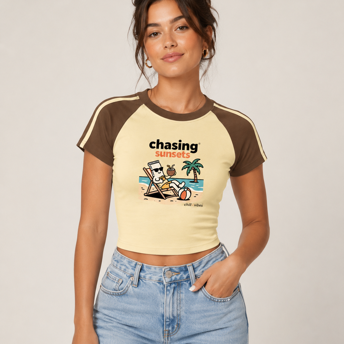 A woman modeling a short-sleeve crop top with brown raglan sleeves and a cream body, featuring a 'chasing sunsets' graphic on the front. She is wearing light-wash blue jeans against a plain light beige studio background.