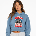 A woman wearing a light blue cropped hoodie with a distressed red, white, and blue graphic on the front that reads 'FREEDOM REBEL ENERGY' and 'Rebel Wear,' paired with light gray sweatpants, against a plain white background.