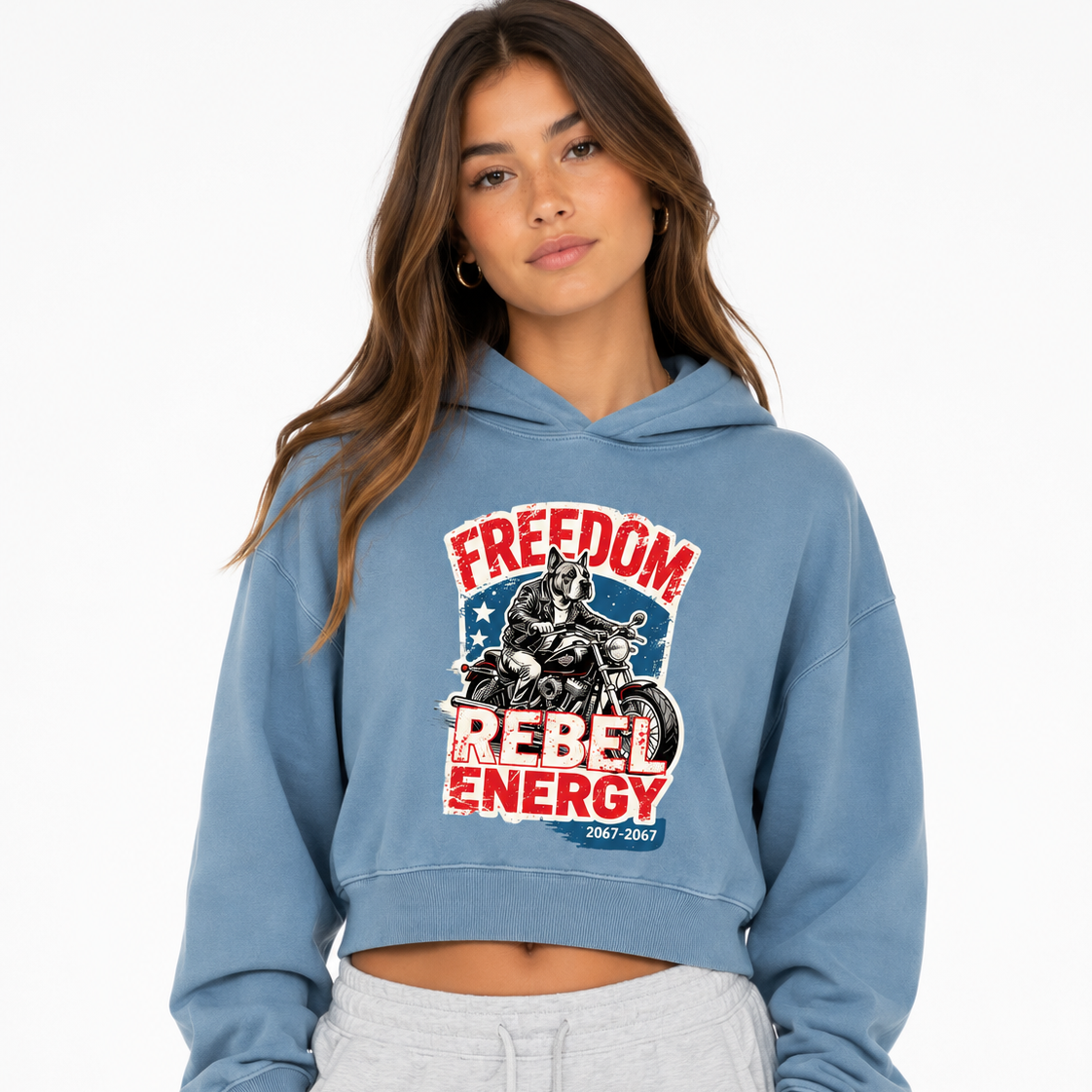 A woman wearing a light blue cropped hoodie with a distressed red, white, and blue graphic on the front that reads 'FREEDOM REBEL ENERGY' and 'Rebel Wear,' paired with light gray sweatpants, against a plain white background.