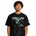 A man wearing a black graphic t-shirt with a colorful hooded character print and the text 'STAY LOW' across the chest, paired with black sweatpants against a plain white background.