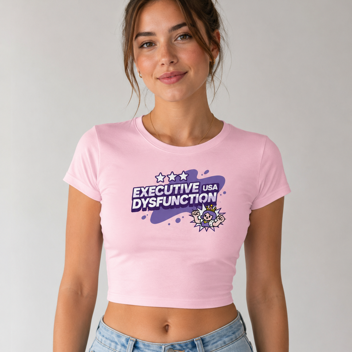 A woman wearing a light pink crop top t-shirt with a purple and white graphic that reads 'EXECUTIVE USA DYSFUNCTION' and a cartoon character, paired with light blue jeans, against a plain light gray background.
