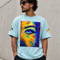 A man wearing a light blue t-shirt with a large, vibrant, multicolored abstract face graphic on the front, set against a neutral concrete wall.